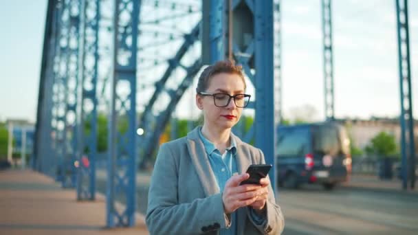 Nő egy kabát sétál a városban kora reggel, és használja okostelefon.. Kommunikáció, munkanap, forgalmas élet koncepció. — Stock videók