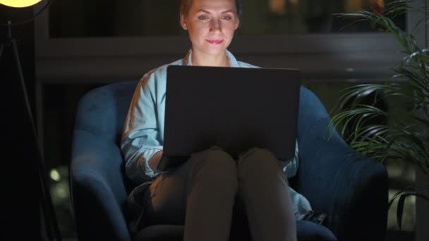 女性はアームチェアに座って、夜にラップトップで作業したり、誰かにメールを送ったりしています。リモートワークの概念. — ストック動画