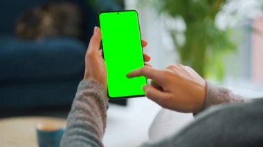 Evde dikey modda yeşil ekranlı akıllı telefon kullanan bir kadın var. Kız internette geziniyor, içeriği izliyor, videolar izliyor. POV.