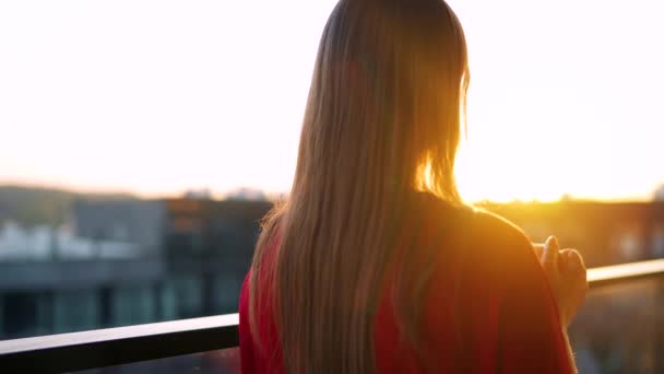 Женщина пьет кофе, стоя на балконе и любуясь закатом. Медленное движение — стоковое видео