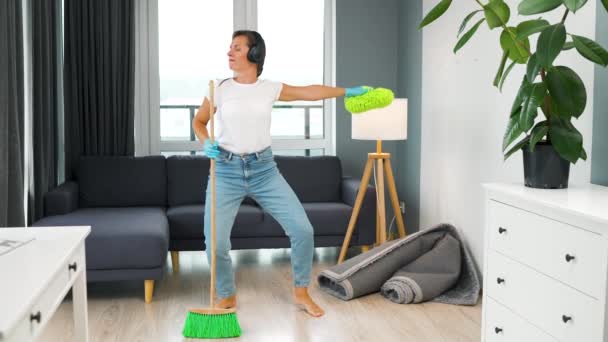 Kvinna i hörlurar städa huset och ha kul dansa med en kvast och tvättlapp. Långsamma rörelser — Stockvideo