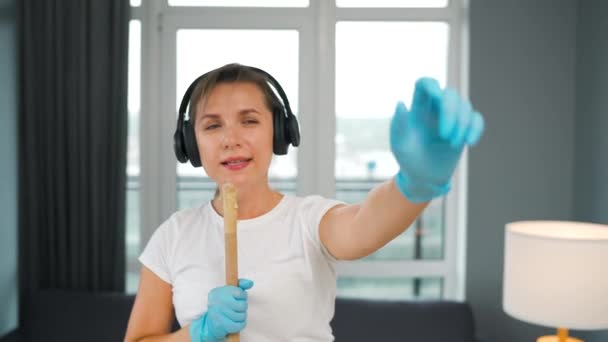 Woman in headphones cleans the house and have fun singing with a broom like a star at a concert. Slow motion — Stock Video