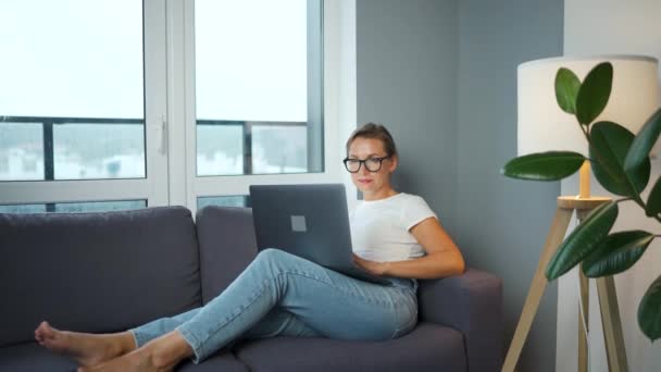 Mulher com óculos está sentada no sofá e trabalhando em um laptop. Conceito de trabalho remoto. — Vídeo de Stock