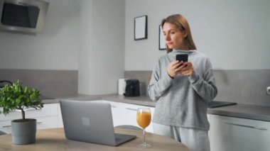 Elinde akıllı telefonu olan bir kadın mutfakta duruyor ve dizüstü bilgisayarda çalışıyor. Serbest çalışıyor, biriyle sohbet ediyor, e-postalara cevap veriyor. Uzak çalışma kavramı.