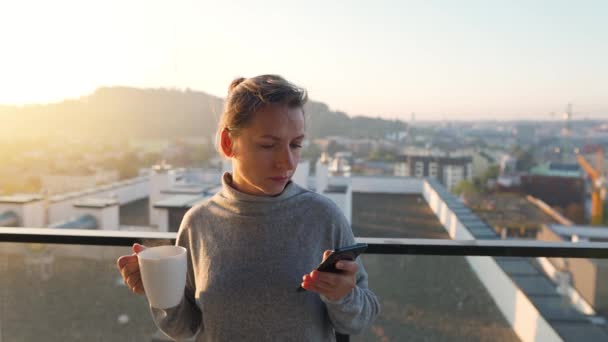 Femme commence sa journée avec une tasse de thé ou de café et vérifier les e-mails dans son smartphone sur le balcon à l'aube, au ralenti. — Video