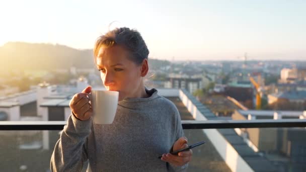 Vrouw begint haar dag met een kopje thee of koffie en het controleren van e-mails in haar smartphone op het balkon bij dageraad, slow motion. — Stockvideo