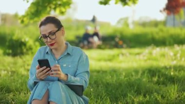 Gün batımında açık havada işi bittikten sonra parkta otururken akıllı telefon kullanan bir kadın. Dışarıda çalışan serbest çalışan biri.