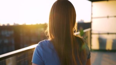 Mavi elbiseli kadın balkonda durup günbatımını seyrederken kahve ya da çay içiyor..