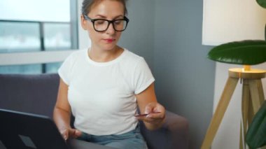Gözlüklü bir kadın rahat bir odada kanepeye oturur ve kredi kartı ve laptop kullanarak online alışveriş yapar. İnternetten alışveriş, yaşam tarzı teknolojisi.