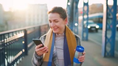 Beyaz ceketli bir iş kadını, soğuk güneşli bir sabahta köprüde yürüyor, kahve içiyor ve akıllı telefon kullanıyor. İletişim, iş günü, modern yoğun yaşam konsepti.