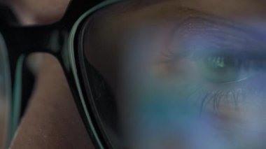 Gözlüklü kadın monitöre bakıyor ve geceleri internette sörf yapıyor. Monitör ekranı gözlüklere yansıyor. Gece çalış. Uzaktan kumanda. Aşırı yakın çekim