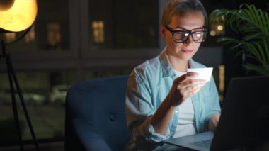 Kadın koltukta oturuyor ve geceleri kredi kartı ve dizüstü bilgisayar kullanarak online alışveriş yapıyor. İnternetten alışveriş, yaşam tarzı teknolojisi.