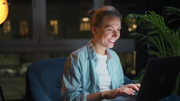 女性はアームチェアに座って 夜にラップトップで作業したり 誰かにメールを送ったりしています リモートワークの概念 内務省 — ストック動画