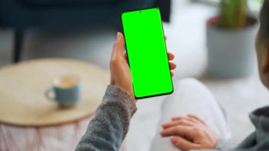Evde dikey modda yeşil ekranlı akıllı telefon kullanan bir kadın var. Kız internette geziniyor, içeriği, videoları, blogları izliyor.