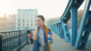 Beyaz ceketli bir iş kadını, soğuk güneşli bir sabahta köprüde yürüyor, kahve içiyor ve akıllı telefon kullanıyor. Ağır çekim. İletişim, iş günü, modern yoğun yaşam konsepti.