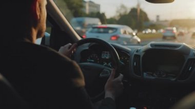 Adam yol boyunca araba sürüyor, batan güneş ön camda parlıyor. Şehir trafiği, arabalar, trafik ışıkları. Sürücülerin omzunun arkasından bak.