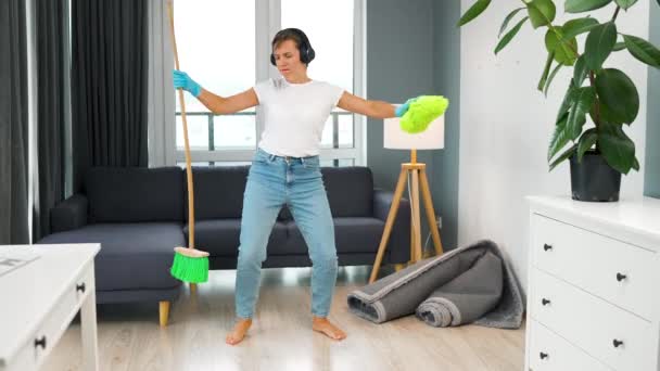 ヘッドフォンの白人女性は家を掃除し ほうきと洗濯布で楽しいダンスをしています — ストック動画