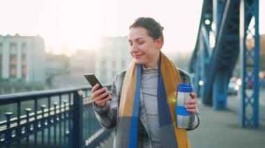 Beyaz ceketli bir iş kadını, soğuk güneşli bir sabahta köprüde yürüyor, kahve içiyor ve akıllı telefon kullanıyor. Ağır çekim. İletişim, iş günü, modern yoğun yaşam konsepti.