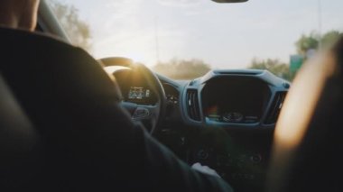 Adam yol boyunca araba sürüyor, batan güneş ön camda parlıyor..