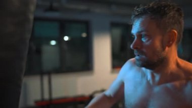 Alone male boxer is working out blows on a punching bag in a gym in evening. Slow motion