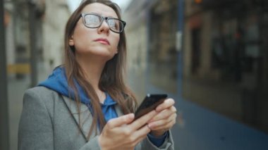 Kadın toplu taşıma durağında duruyor ve zaman çizelgesini kontrol ediyor. Tramvay arka planda duruyor. Şehir, şehir, ulaşım.