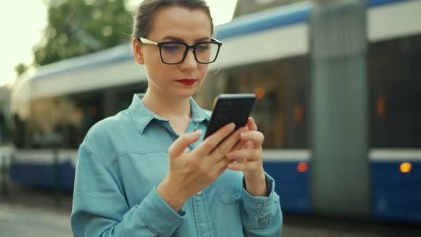 Woman Stands Public Transport Stop Using Smartphone Tram Pulls Background — Vídeo de stock