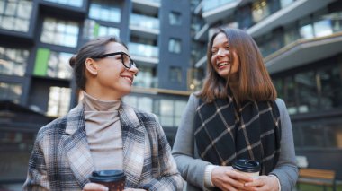 İki mutlu kadın hazır kahveyle yürüyüp kendi aralarında faiziyle konuşurlar. Arka plandaki modern binalar