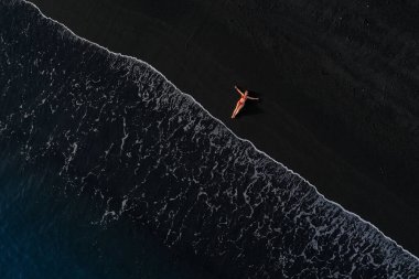 Sörf hattında siyah bir plajda uzanan kırmızı mayo giymiş bir kızın hava görüntüsü. Tenerife adasının kıyıları, Kanarya Adaları, İspanya.