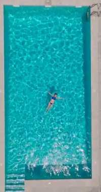Vertical video. Top down view of a woman in a blue swimsuit lying on her back in the pool. Relaxing concept, summer lifestyle