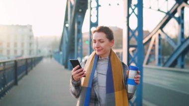 Caucasian businesswoman in a coat, standing on the bridge in a frosty sunny morning, drinking coffee and using smartphone. Slow motion. Communication, work day, modern busy life concept.