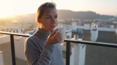 Kadın güne şafakta balkonda bir fincan çay ya da kahveyle başlıyor, ağır çekimde. Arka planda şehir manzarası var. Modern şehir yaşam tarzı
