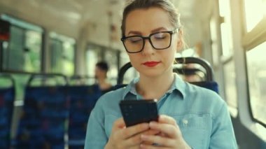 Public transport. Slow motion of woman in glasses in tram using smartphone chatting and texting with friends. City, urban, transportation.