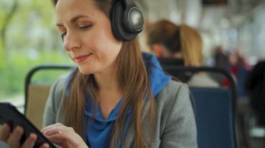 Public transport. Woman in headphones in tram using smartphone chatting and texting with friends. City, urban, transportation.