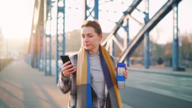 Paltolu beyaz bir iş kadınının portresi, soğuk güneşli bir sabahta şehirde geziniyor, kahve içiyor ve akıllı telefon kullanıyor. İletişim, iş günü, yoğun yaşam konsepti. Yavaş çekim