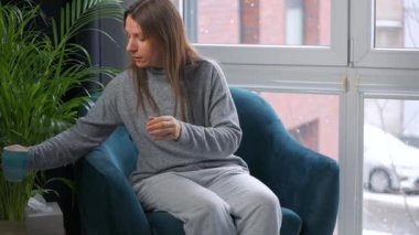 Unhealthy caucasian woman sits in a chair, drinks hot tea or a cold medicine and coughs into a napkin because she has a cold, flu, coronavirus. It is snowing outside