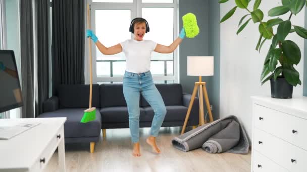 ヘッドフォンで白人女性は家を掃除し ほうきや洗濯で楽しいダンスをしています スローモーション — ストック動画