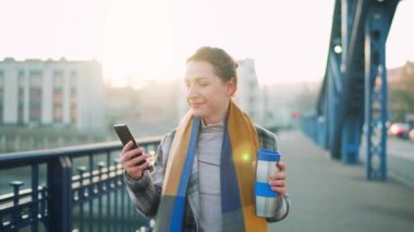 Beyaz ceketli bir iş kadını, soğuk güneşli bir sabahta köprüde yürüyor, kahve içiyor ve akıllı telefon kullanıyor, ağır çekimde. İletişim, iş günü, modern yoğun yaşam konsepti.