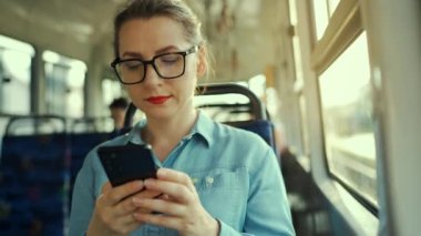 Public transport. Slow motion of woman in glasses in tram using smartphone chatting and texting with friends. City, urban, transportation.