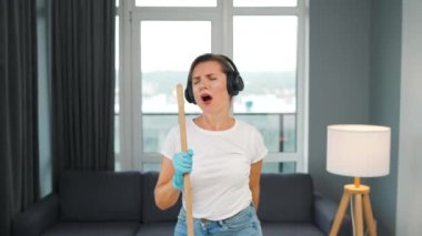 Beyaz kulaklıklı kadın evi temizliyor ve konserdeki bir yıldız gibi süpürgeyle şarkı söyleyerek eğleniyor. Yavaş çekim