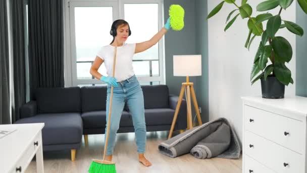ヘッドフォンの白人女性は家を掃除し ほうきと洗濯布で楽しいダンスをしています — ストック動画