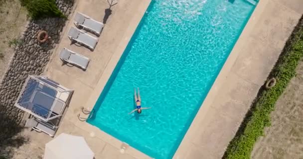一个穿着蓝色泳衣的女人躺在游泳池里的自上而下的照片 相机缓慢地逆时针旋转 并在游泳池上方下降 放松概念 夏季生活方式 — 图库视频影像