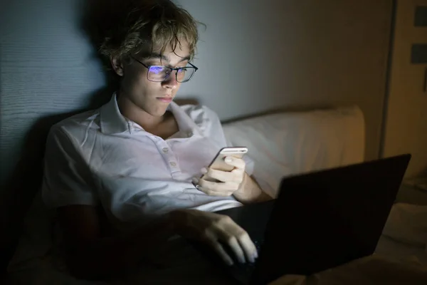 一个年轻人躺在笔记本电脑前 手里拿着智能手机 躺在床上 青少年晚上做作业或与朋友交流 — 图库照片