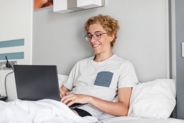 Gülümseyen, 20 'li yaşlarda gri tişört giyen, dizüstü bilgisayarla çalışan, çizgili çarşaf yastık battaniyesiyle yatakta uzanan, yatak odasında dinlenen, gevşetici bir adam. Kampta dizüstü bilgisayarı olan mutlu genç çocuk.