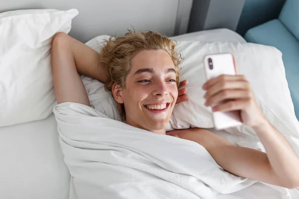 随意微笑的年轻人在家里的床上发短信电话 早上快乐英俊的男人看手机 — 图库照片