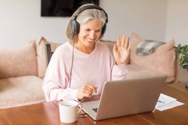 Kulaklıklı mutlu orta yaşlı kurs öğrencisi dizüstü bilgisayarda Webinar izlemeyi öğreniyor. Olgun bir kadın internette yeni beceriler öğreniyor, notlar alıyor. İş kadını görüntülü görüşme yapıyor ve elini evden sallıyor.