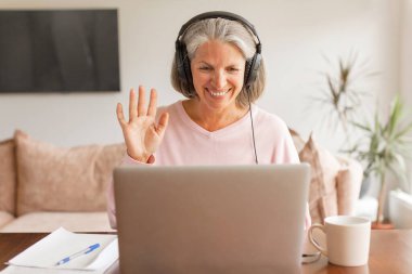 Mutlu 50 'li yaşlarda, kulaklıklı, gri saçlı, merhaba jesti yapan, bilgisayar uygulaması, yaşlı insanlar ve modern teknoloji uzaktan iletişimi kullanarak online video görüşmeleri başlatan,.