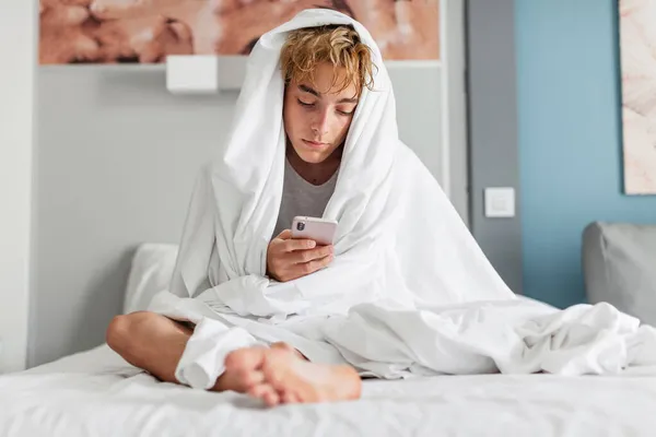 一个孤独而又忧心忡忡的男人坐在床上看着手机等着电话 在卧室里拿着智能手机的可悲的青少年 — 图库照片