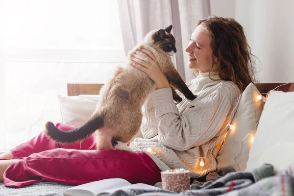 穏やかな女性は窓の近くのソファでリラックスし ふわふわの猫を撫でています 彼女は動物を見ている — ストック写真