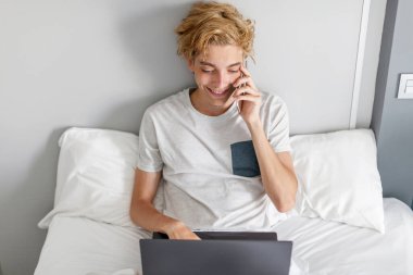 Evde yatağında dinlenirken cep telefonu kullanan, dizüstü bilgisayarlı, beyaz, genç bir adam. Evde yatak odasında telefonu ve bilgisayarı olan bir çocuk.