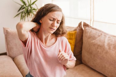 Yorgun genç bir kadın boynuna dokunuyor eklem ağrısı acıyor masaj yapıyor gergin kaslar fibromiyaljiden muzdarip uzun bilgisayar çalışmalarının ardından yanlış bir duruşla evdeki koltukta oturuyor.